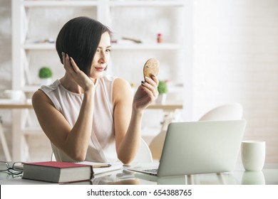 Beautiful Young Woman Looking In Small Mirror At Workplace With Laptop, Coffee Cup And Book. Self Obsession Concept