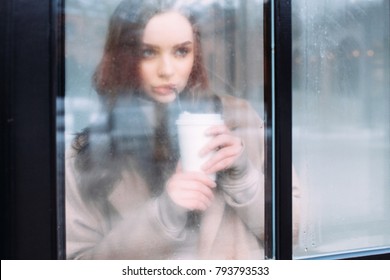 Beautiful young woman are looking thru the window in winter and drinks coffee alone