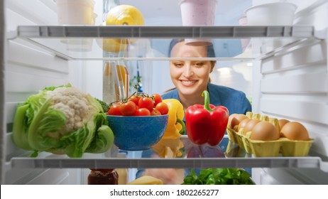 Beautiful Young Woman Looks Inside The Fridge And Takes Out Vegetables. Woman Preparing Healthy Meal Using Groceries. Point Of View POV From Inside Of The Kitchen Refrigerator Full Of Healthy Food