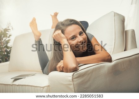 Beautiful young woman lying face down on a white living room couch with forearms resting on the armrest and legs bent behind with bare feet up, looking super relaxed and happy, cheerful like a child -