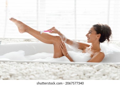 Beautiful young woman lying in foamy bath, shaving her legs with razor at home. Lovely millennial lady enjoying domestic spa procedure, making depilation in bathtub at luxury hotel