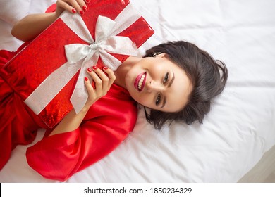 Beautiful Young Woman Lying On The Bed With Red Gift And Smiling. Sexy Woman In Red Dressing Gown With Christmas, Valentine Day Or Birthday Gift