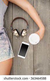 Beautiful Young Woman Lying On The Wooden Floor With Music Headphones, Smartphone And A Take Away Coffee Cup; View From Above.