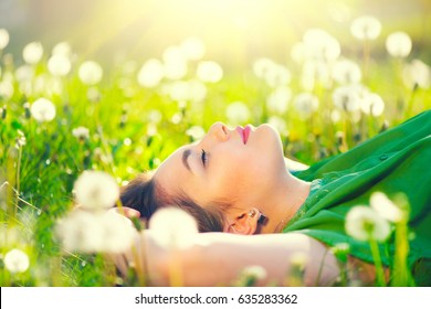 Beautiful Young Woman lying on the field in green grass and dandelions and smiling. Outdoors. Enjoy Nature. Healthy Smiling Girl on spring lawn. Allergy free concept