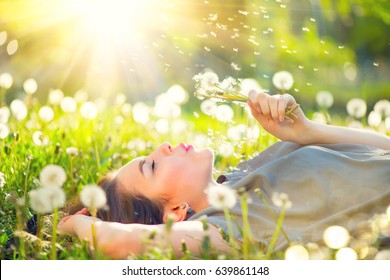 Beautiful Young Woman lying on the field in green grass and blowing dandelion. Outdoors. Enjoy Nature. Healthy Smiling Girl on spring lawn. Allergy free concept. Freedom - Powered by Shutterstock