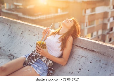 Beautiful young woman lying on the building rooftop edge, drinking pineapple cocktail and enjoying amazing summer sunset