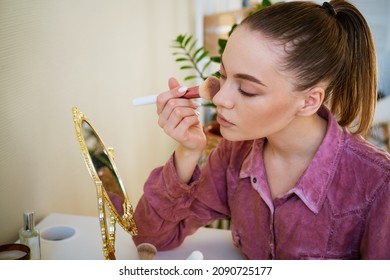 Beautiful Young Woman Makes Blush On Her Face Using Makeup Brush While Sitting At Home In A Room At A Table. Caucasian Girl In A Dark Pink Shirt Does Makeup On Her Own.