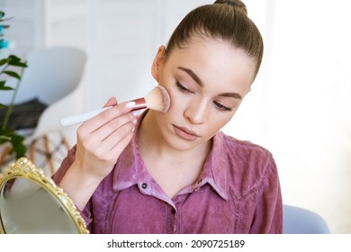 Beautiful young woman makes blush on her face using makeup brush while sitting at home in a room at a table. Caucasian girl in a dark pink shirt does makeup on her own.