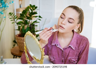 Beautiful Young Woman Makes Blush On Her Face Using Makeup Brush While Sitting At Home In A Room At A Table. Caucasian Girl In A Dark Pink Shirt Does Makeup On Her Own.