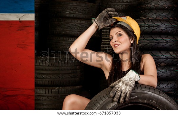 Beautiful young woman mechanic with helmet and\
protective gloves at work in auto service station. Concept works\
with tires on wheels