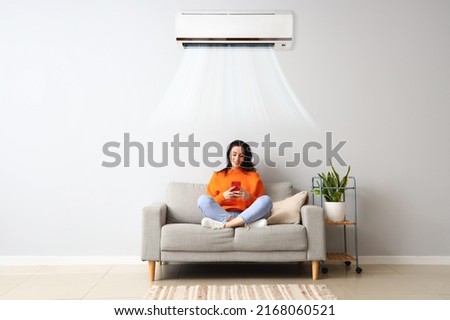 Beautiful young woman with mobile phone resting on sofa in light room with operating air conditioner