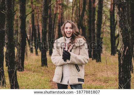 
Beautiful young woman model appearance with long curly hair holding a Christmas candy in the form of a heart and smiling while being in a pine forest