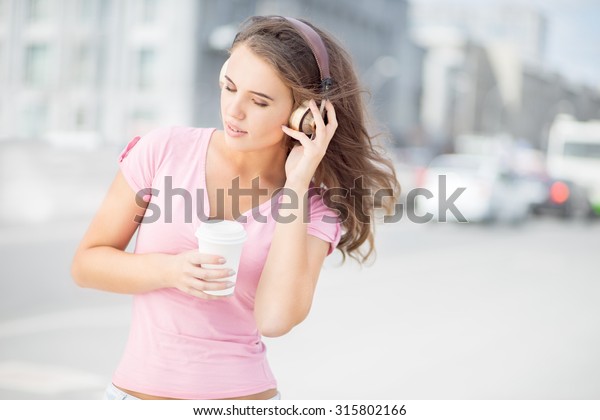 Beautiful young woman with music\
headphones, holding a take away coffee cup and listening to the\
music with her eyes closed against city traffic\
background.