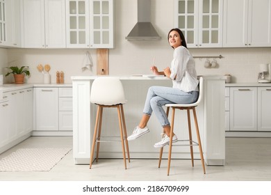 Beautiful young woman with notebook sitting on stool in kitchen - Shutterstock ID 2186709699