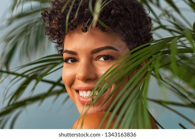 Beautiful young woman with nude makeup looking trought palm leaves. Brazilian smiling woman looking at camera with palm leaves near face. Closeup face of happy african girl in the nature.