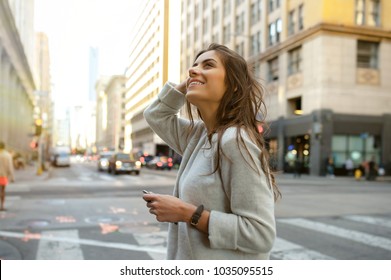 Beautiful young woman on the boulevard in urban scenery, downtown, at sunset, holding smartphone and looking side.