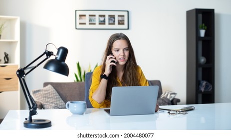 Beautiful Young Woman On A Cell Phone Call Feeling Focused While Working On Laptop In A Light Modern Interior Livingroom Sitting Behind A Big White Desk With Notes.