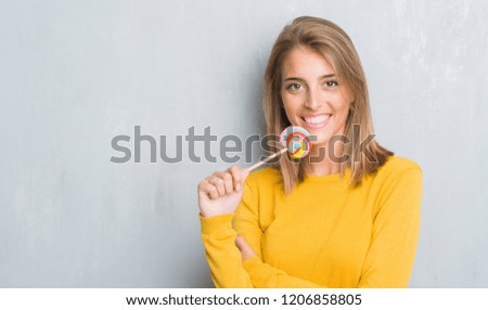 Beautiful young woman over grunge grey wall eating lollipop candy with a happy face standing and smiling with a confident smile showing teeth