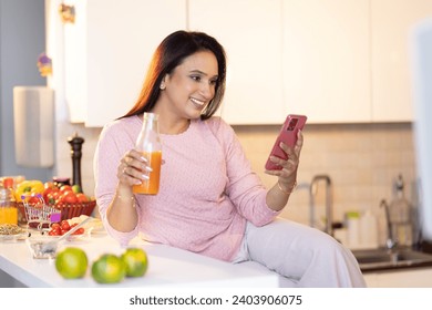 Beautiful young woman in pajamas sitting on kitchen counter, drinking juice and talking on phone with friend - Powered by Shutterstock