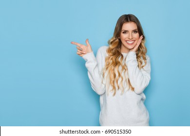 Beautiful Young Woman In Pastel Blue Sweater Is Holding Hand On Chin, Looking At Camera, Pointing And Smiling. Waist Up Length Studio Shot On Turquoise Background.