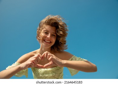 Beautiful Young Woman With Perfect Skin Smiling To The Camera And Shows Heart By Hands. Portrait Of Attractive Woman With Nice Smile. Freedom, Love And Summer Vacation Concept.