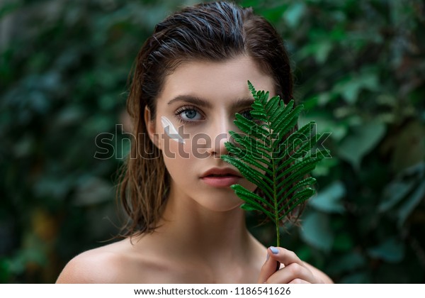 Beautiful young woman with perfect skin and
natural make up posing front of plant tropical green leaves
background with fern. Teen model with wet hair care of her face and
body. SPA, wellness