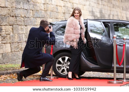 Beautiful young woman and photographer near car on red carpet, outdoors