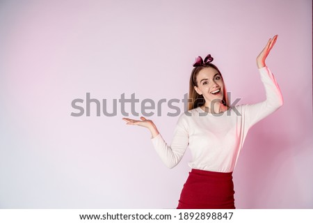 A beautiful young woman in a pink blouse and crimson skirt spreads her hands, looks at the camera and screams. A three-quarter-length studio shot on a pink background with a model.