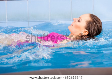 Beautiful young woman in pink swimsuit swimming in blue pool on her back. Young female swimmer at holiday resort. Sport activity health concept.