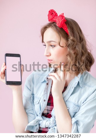 Beautiful young woman with pin-up make-up and hairstyle over pink background with mobile phone with copy space.