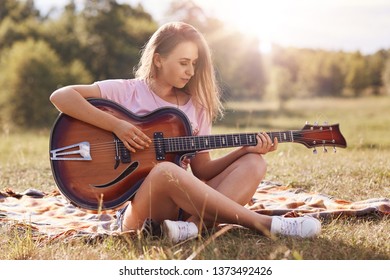 Beautiful Young Woman Playing Gitar In Meadow, Has Stright Blonde Hair, Looks Concentrated On Musical Insrtument, Wearing Casual Clothes, Spending Time Alone, Enjoying Wonderful Nature. Outdoor Shot