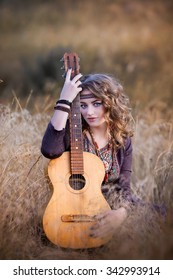 Beautiful Young Woman Playing Gitar On The Field. Perfect Skin And Make-up. Blond Curly Hair. Outdoor Shot.