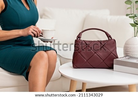 A beautiful young woman poses in a green dress against the background of a sofa in the living room. A woman is holding a burgundy shoulder bag made of braided leather. The concept of women's fashion.