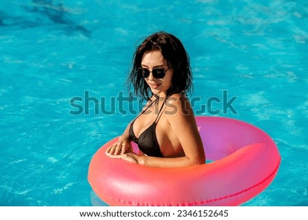 
Beautiful, young woman posing in the pool with a pink inflatable ring. Happy woman on vacation on a warm summer day.