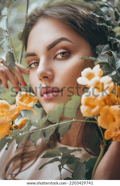 Beautiful young woman posing in Trumpet vine\
flowers in summer garden. Beauty model girl with Campsis. Enjoying\
nature outdoor. Close up\
portrait