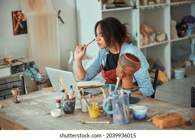 Beautiful young woman potter using laptop in pottery workshop