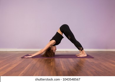 Beautiful Young Woman Practicing Yoga Indoors. Wearing Black Sportswear. Using Purple Mat. Wood Floor And Violet Background. Sports And Lifestyle