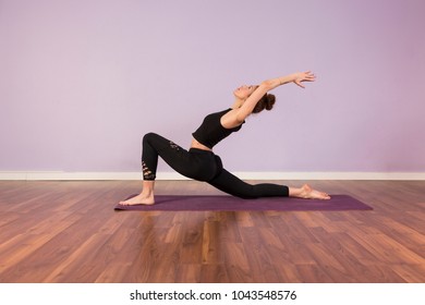 Beautiful Young Woman Practicing Yoga Indoors. Wearing Black Sportswear. Using Purple Mat. Wood Floor And Violet Background. Sports And Lifestyle