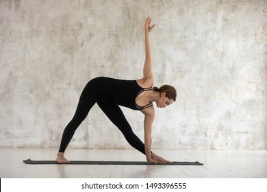 Beautiful young woman practicing yoga, doing extended triangle exercise, girl wearing black sportswear standing in Utthita Trikonasana pose on mat, working out at home or in modern yoga studio