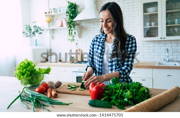Beautiful young woman is preparing vegetable salad\
in the kitchen. Healthy Food. Vegan Salad. Diet. Dieting Concept.\
Healthy Lifestyle. Cooking At Home. Prepare Food. Cutting\
ingredients on table