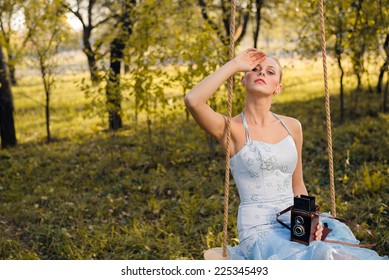 Beautiful Young Woman In Prom Or Cocktail Dress Sitting On Swings With Retro Photo Camera On Green Summer Or Autumn Outdoors Copyspace Background