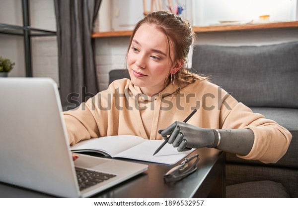 Beautiful young woman with\
prosthesis arm writing at notebook while sitting at the table with\
her laptop computer. Adorable girl looking at the screen while\
working at home