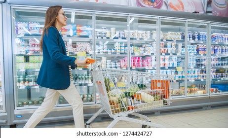 Beautiful Young Woman Pushes Shopping Cart full of Goods Through Frozen Goods and Dairy Section of the Supermarket.