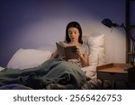 Beautiful young woman reading book on bed in evening at home