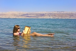Beautiful Young Woman Reads A Book Floating In The Waters Of The Dead Sea In Israel