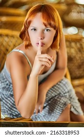 Beautiful young woman with red hair sits in wooden wicker armchair and calls for silence applying index finger to lips.
