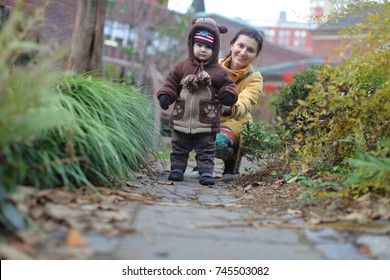 Beautiful young woman relaxing in garden, walking with baby, having fun with family. Female model in yellow coat and colorful jeans. Little baby toddler in knitted coat like a bear. - Powered by Shutterstock