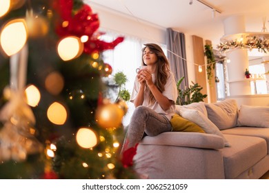 Beautiful young woman relaxing at home on Christmas day, sitting on couch by the Christmas tree, drinking coffee