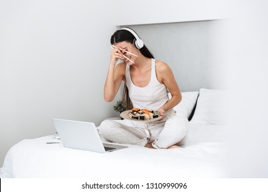 Beautiful Young Woman Relaxing On Bed At Home, Listening To Music With Headphones, Eating Sushi From A Plate