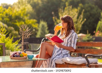 Beautiful Young Woman Relaxing On A Sunny Summer Morning In Her Backyard, Having Breakfast, Drinking Orange Juice And Reading A Book Outdoors